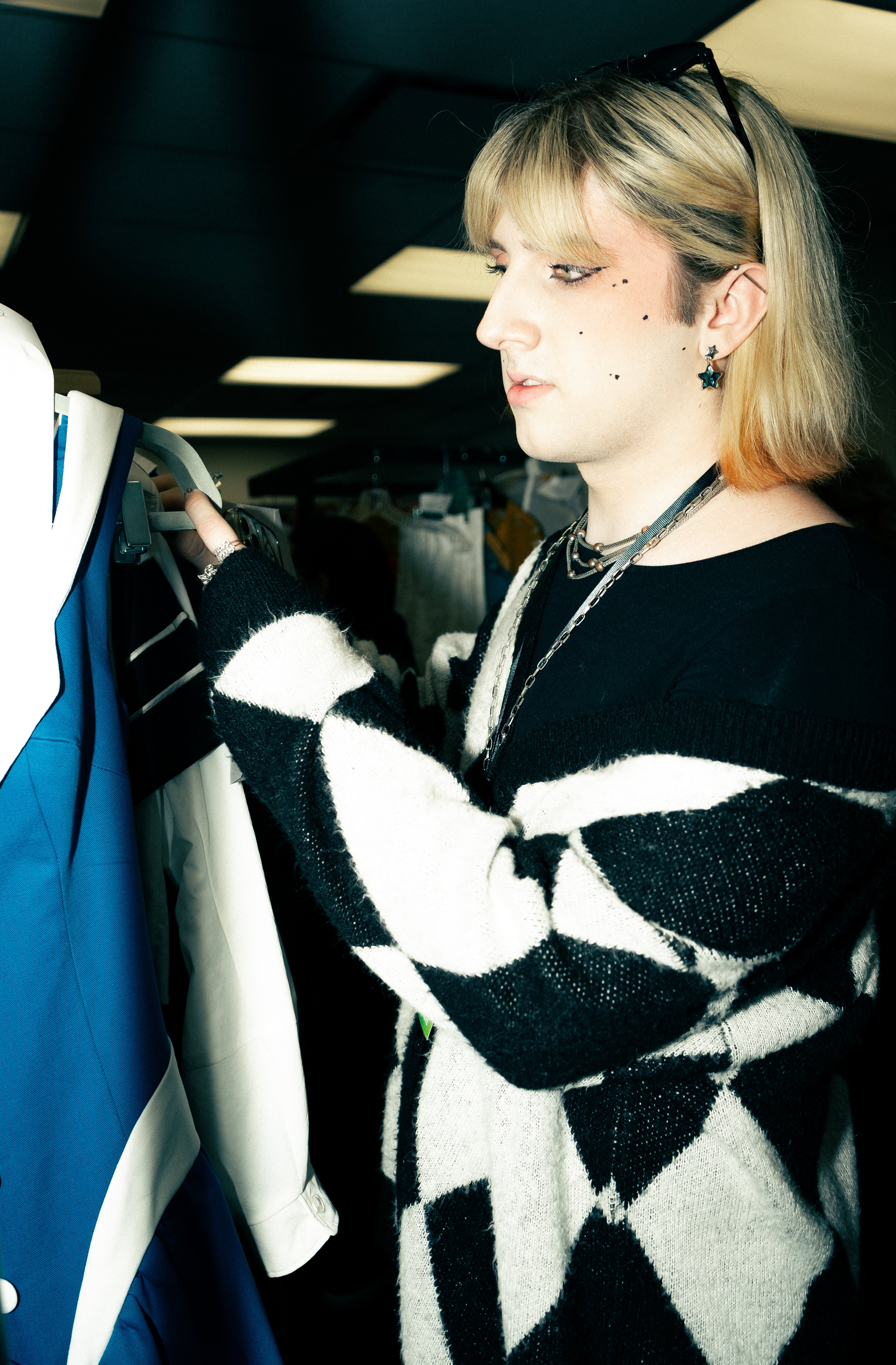 A person examining a blue garment on a hanger, wearing a black and white patterned sweater and multiple necklaces, with short blonde hair styled with bangs.